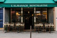 La façade de l’Almanach Montmartre révèle un élégant immeuble haussmannien sublimé par une enseigne discrète et un portillon en fer forgé. Ses fenêtres à petits carreaux et son entrée encadrée de boiseries noires annoncent l’esprit intime et bohème qui rè | © L'Almanach Montmartre